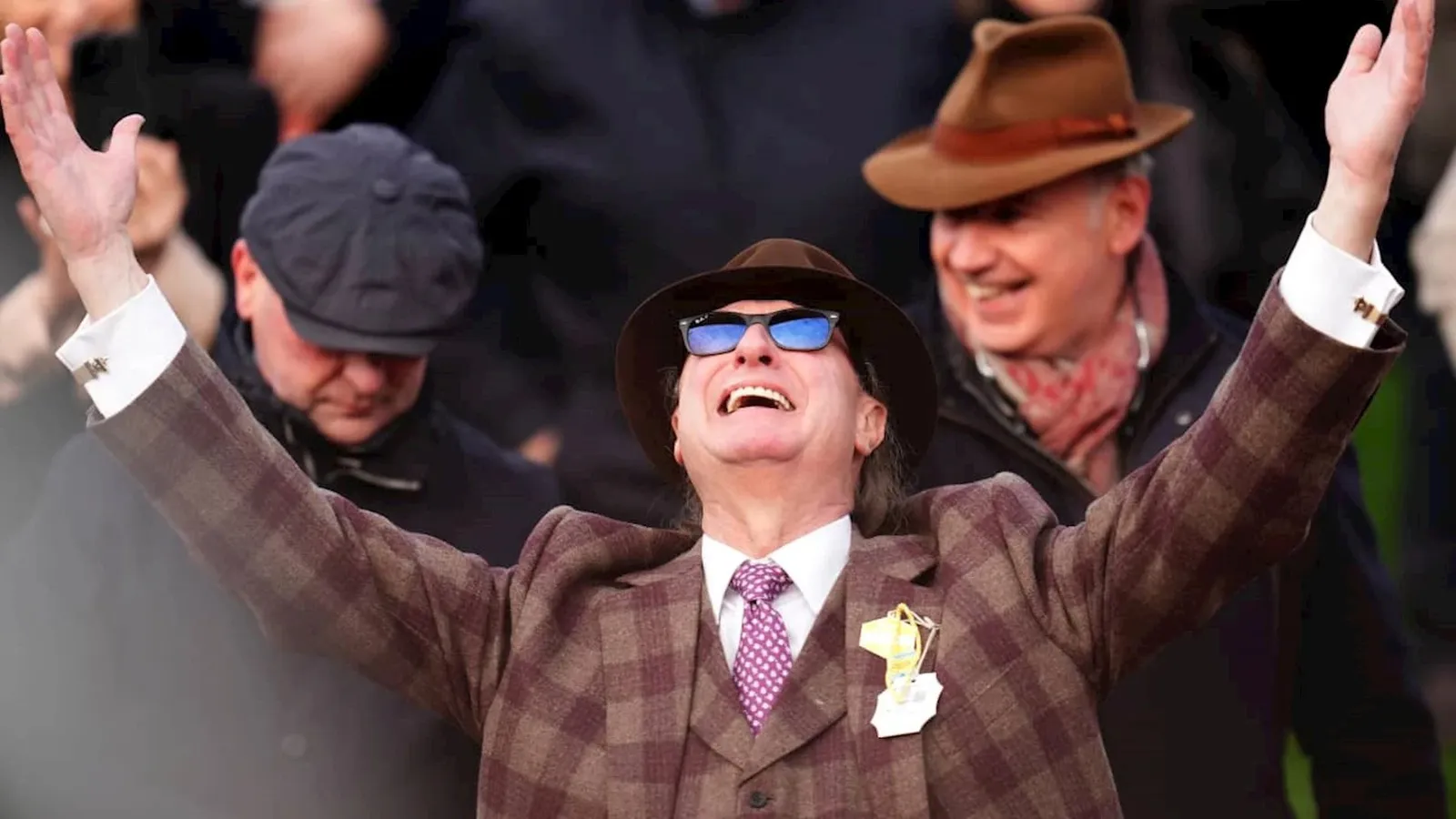 A racegoer in a check tweed three-piece suit, a tie and a trilby cheering with both arms raised at Cheltenham.