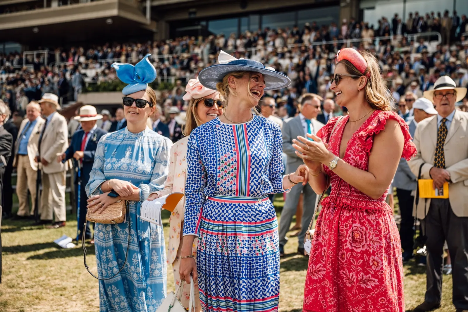 Three women in printed summer dresses and hats laughing on the lawn at Goodwood, with men in linen jackets and Panama hats behind them.