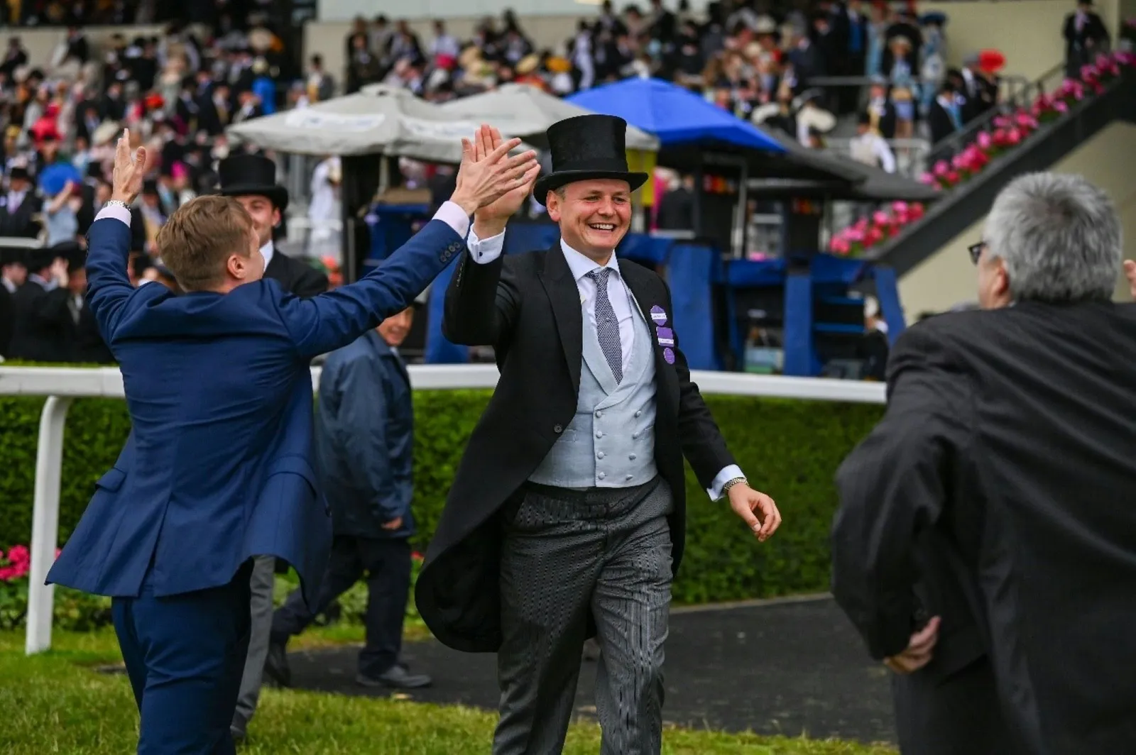 Two men in morning dress and top hats high-fiving beside the rail at Royal Ascot.