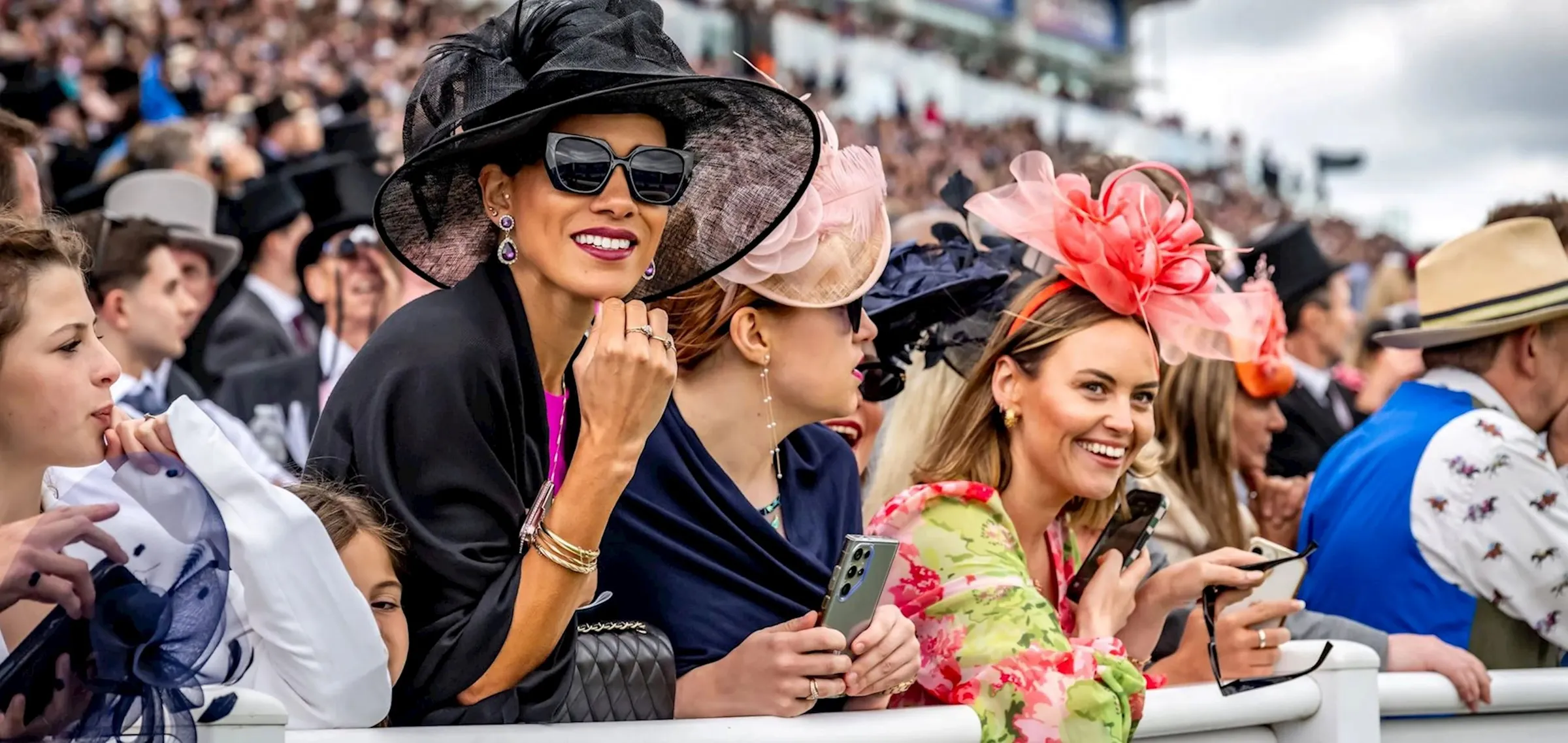 Racegoers along the rail at Epsom in wide-brimmed hats and fascinators, with men in morning dress and top hats behind them.