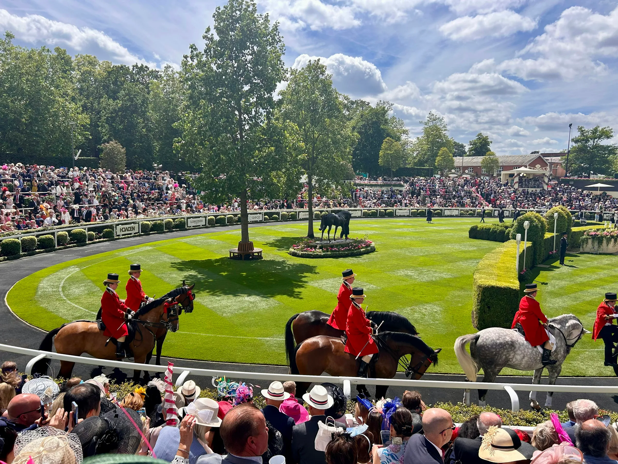 Ceremonial outriders in scarlet on horseback in the parade ring at Royal Ascot, ringed by a hatted crowd under the trees.