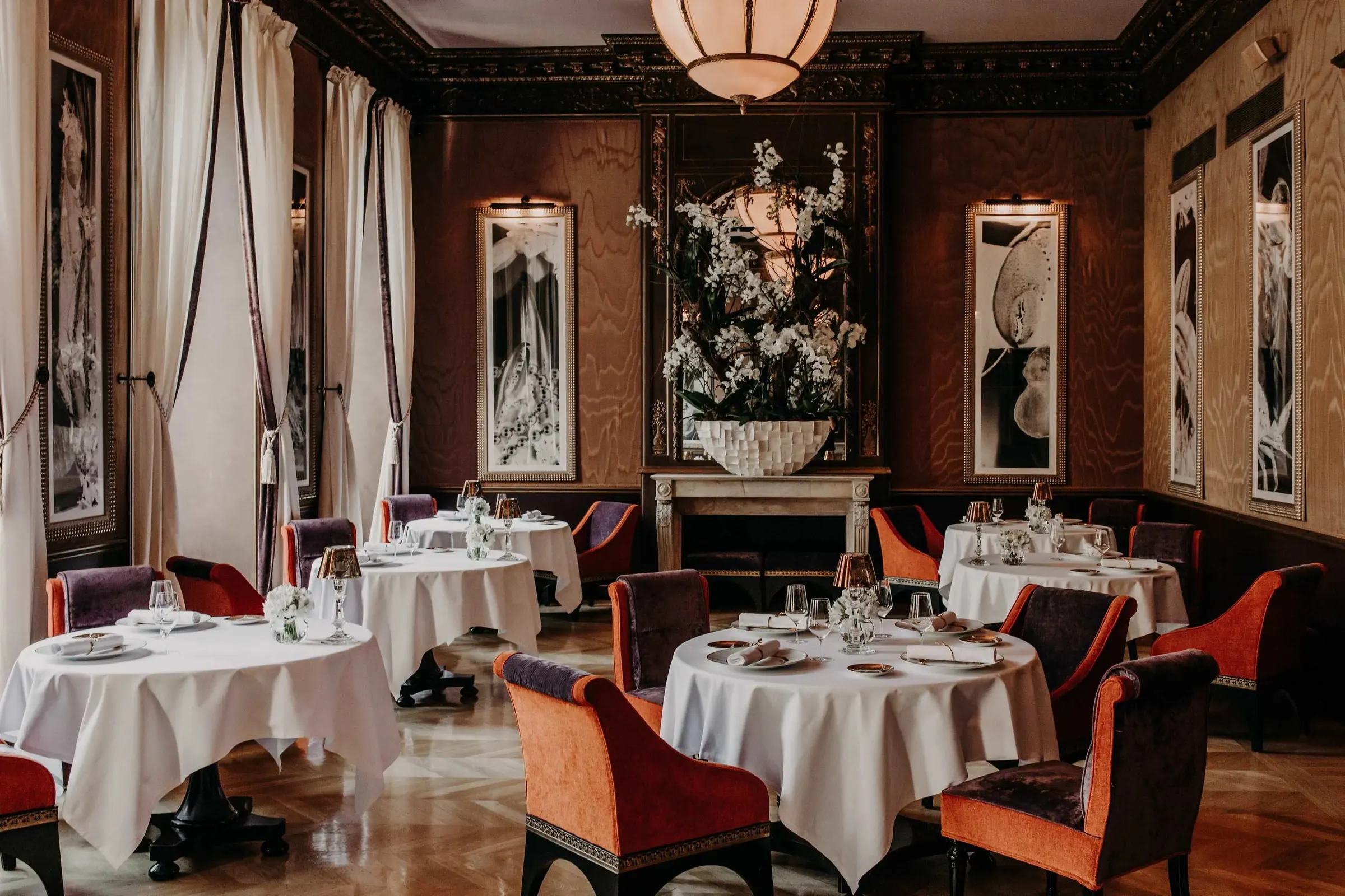 A formal dining room laid for dinner, with white tablecloths, orange and purple velvet chairs and a large white orchid display.