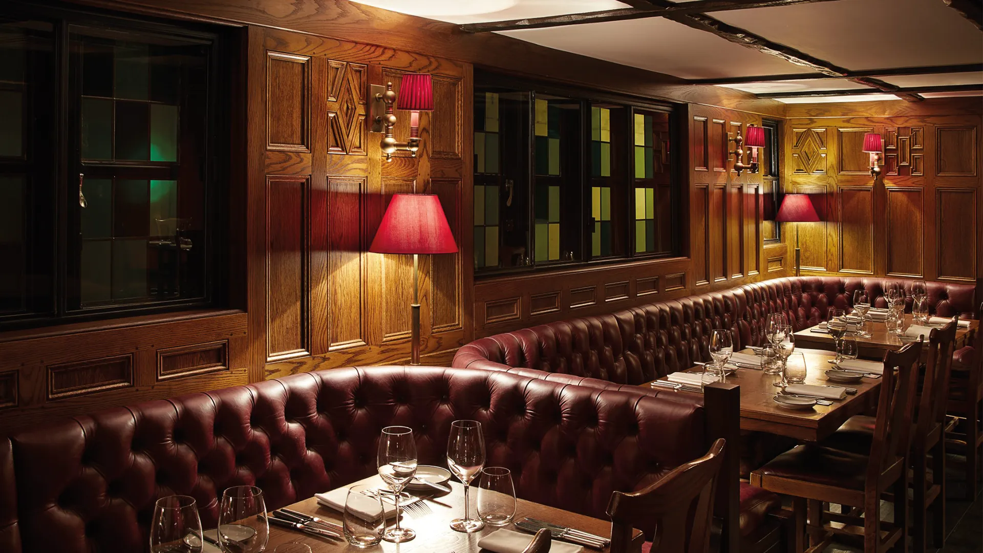 The oak-panelled dining room at The Hind's Head, with button-backed leather banquettes, red-shaded lamps and tables laid with wine glasses.