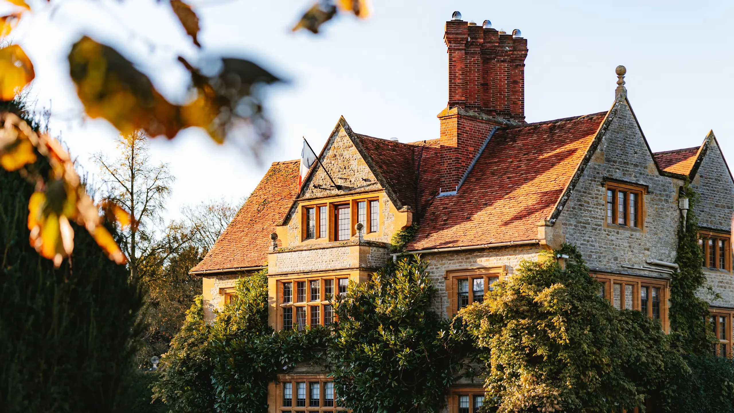The honey-stone manor house of Le Manoir aux Quat'Saisons in low autumn sun, its walls covered in climbing plants above a lawn, with golden leaves in the foreground.
