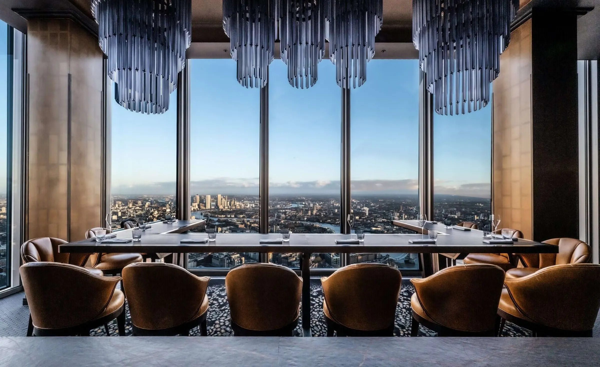A high-rise London dining room with floor-to-ceiling windows over the city and Canary Wharf, a long table under glass chandeliers.