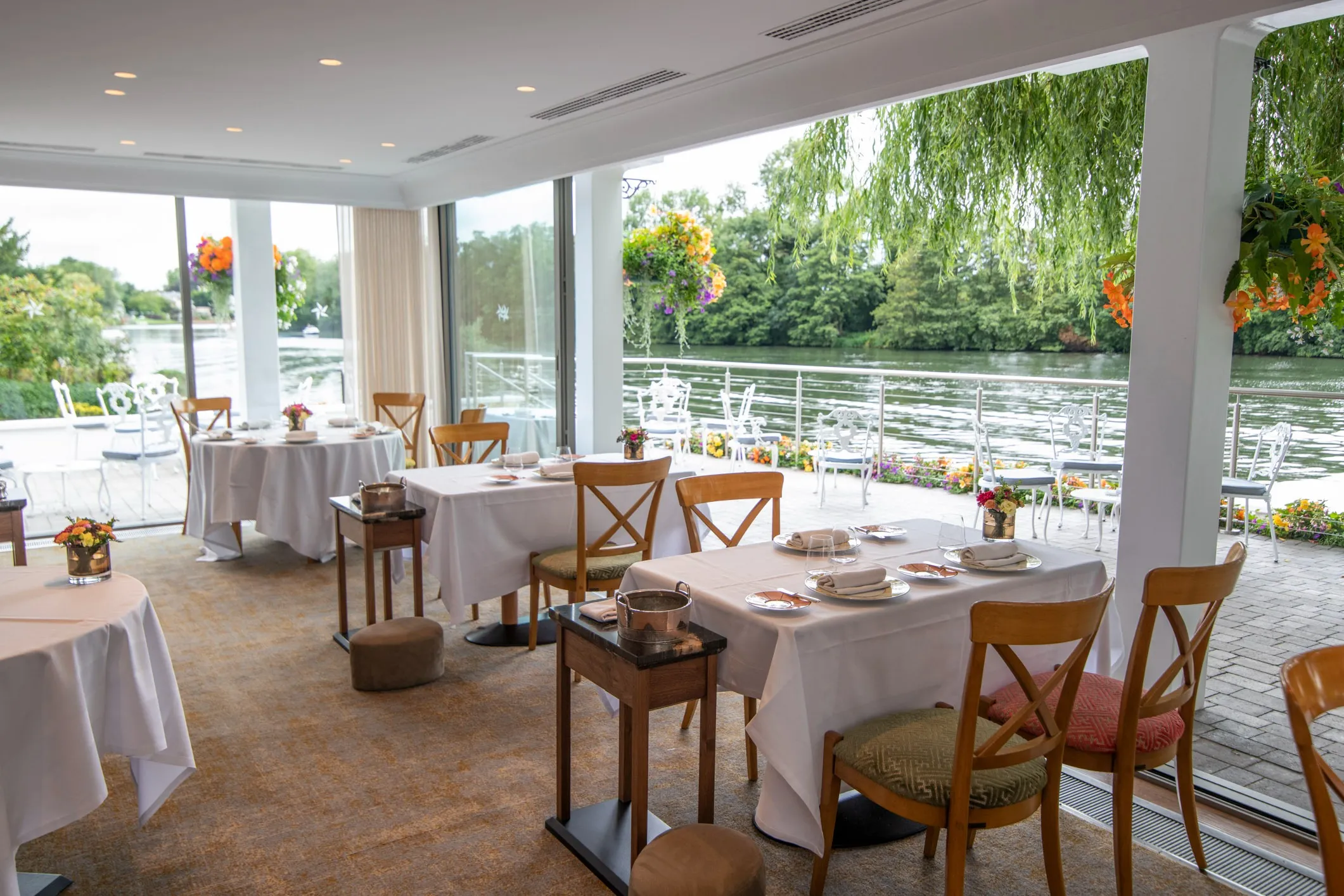 The riverside dining room at The Waterside Inn, its white-clothed tables laid for service beside open glass doors onto a terrace and the Thames.