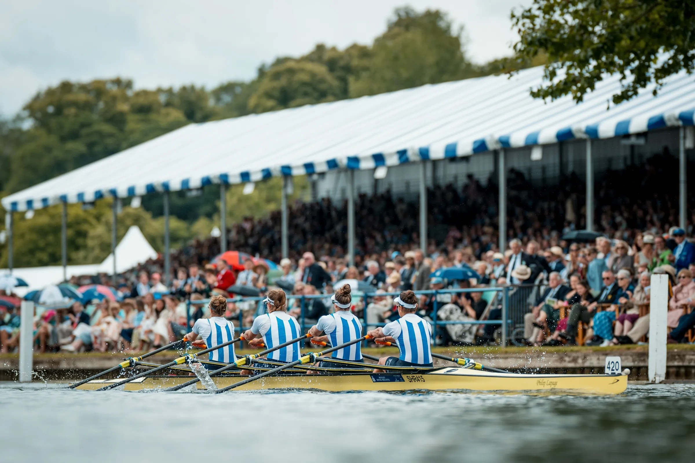 A rowing crew racing past a striped grandstand full of spectators at Henley.