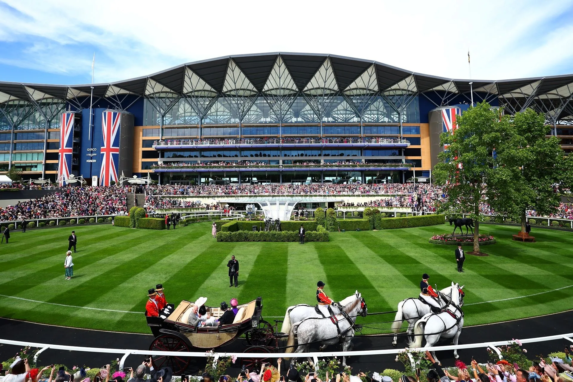 A carriage drawn by grey horses crossing the striped lawn in front of the grandstand at Royal Ascot, with Union Flags hanging from the stand.