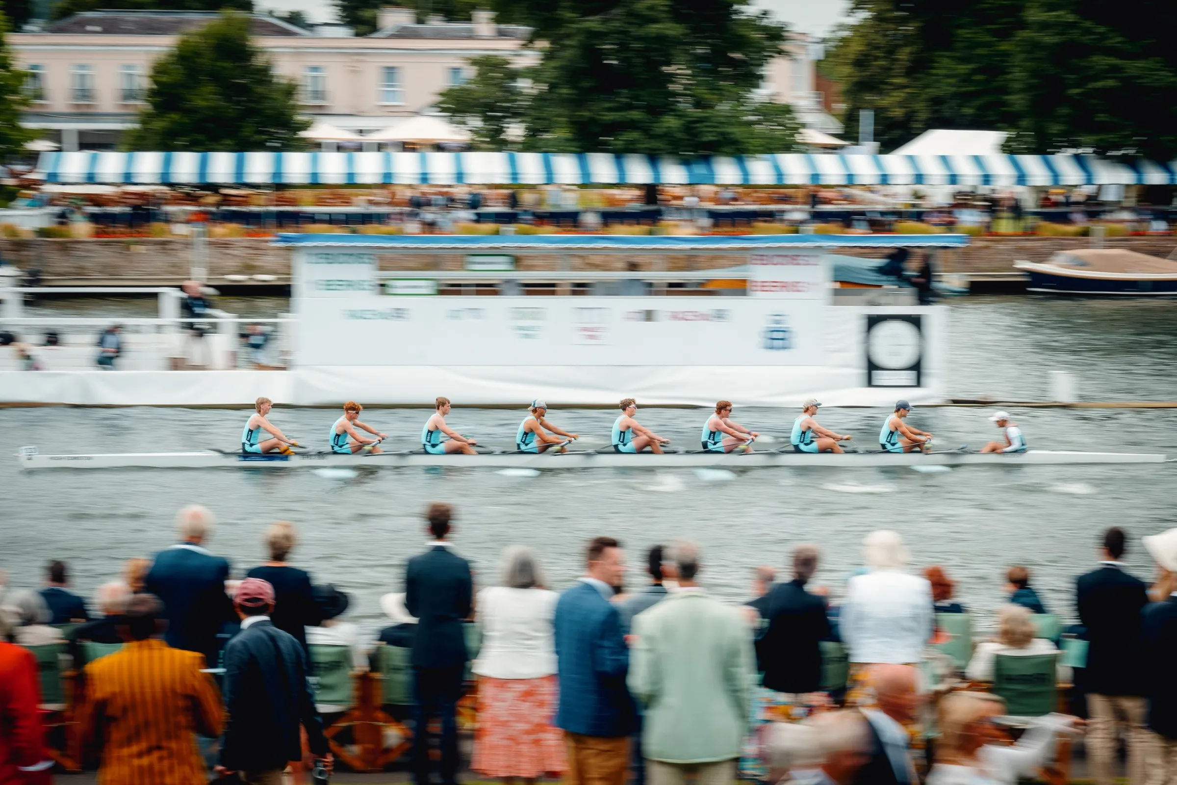 A rowing crew racing past the enclosures at Henley, with spectators on the bank in the foreground.