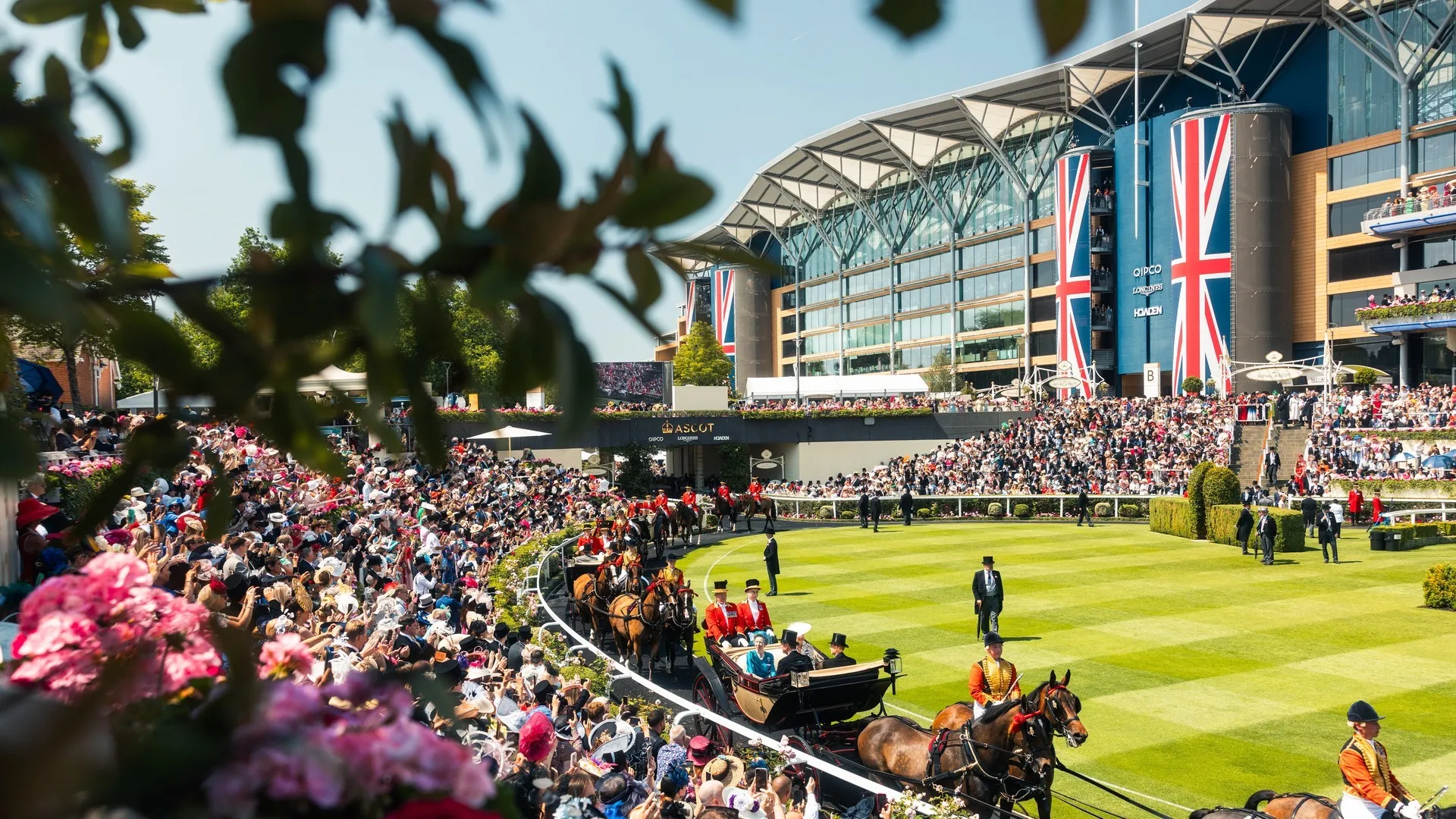Carriages of the Royal Procession crossing the parade ring at Royal Ascot, with Union Flags hanging from the grandstand behind.