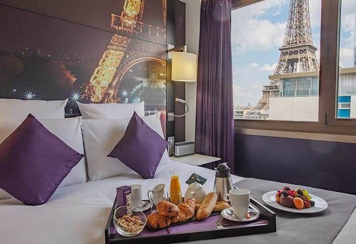 Image of hotel bed with continental breakfast on top with a window view of the effiel tower